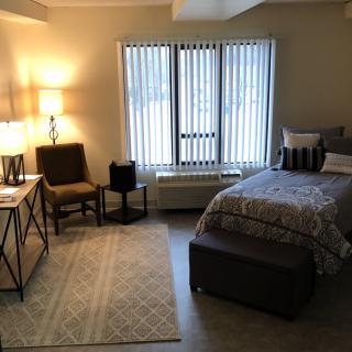 A cozy bedroom with a neatly made bed, a desk with a lamp, and comfortable seating by the window.