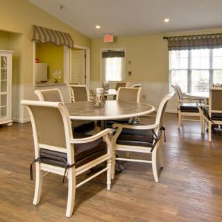 A cozy dining area with a round table and cushioned chairs, featuring large windows and wooden flooring.