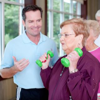 An elderly woman exercises with dumbbells under the guidance of a trainer.