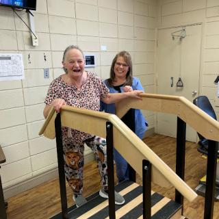 An elderly woman joyfully practices walking up stairs with the assistance of a caregiver.