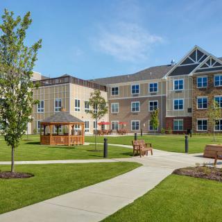 A serene view of a landscaped area with a modern building, pathways, benches, and a gazebo.