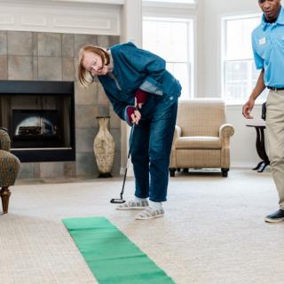 An elderly woman practices putting on an indoor golf green with assistance in a living room setting.