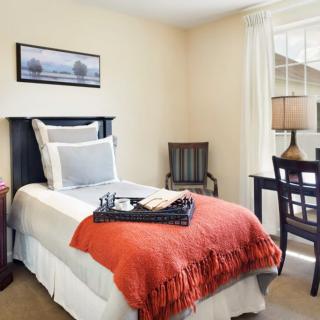A cozy bedroom with a single bed adorned with an orange blanket, featuring a desk and a lamp by the window.