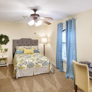 A cozy bedroom with a bed, desk, and ceiling fan, featuring blue curtains and elegant decor.