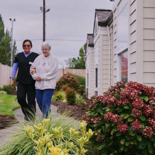 A healthcare worker walks with an elderly person through a garden pathway.