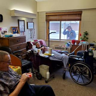 An elderly nursing home resident receives a visitor at the window while another resident sits nearby.
