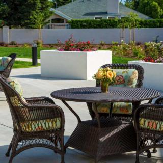 A sunny patio with wicker chairs and a table adorned with flowers, surrounded by a well-maintained garden.