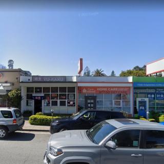 A busy urban street with parked cars in front of storefronts on a sunny day.