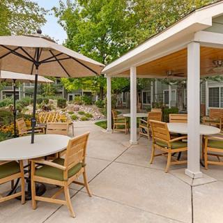 A cozy outdoor patio area with tables and chairs, shaded by umbrellas, set amid a landscaped garden.