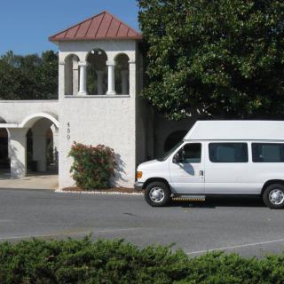 A white van parked in front of a building with a tower and surrounded by trees.