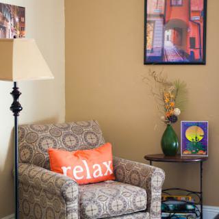 A cozy corner in a living room featuring a patterned armchair with a 'relax' pillow, a floor lamp, and decorative wall art.