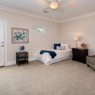 A spacious, neutral-colored bedroom featuring a single bed with blue accents, a nightstand with a lamp, a decorative painting, and a comfortable chair.