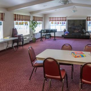 A spacious community room with tables and chairs, a piano, and large windows providing natural light.