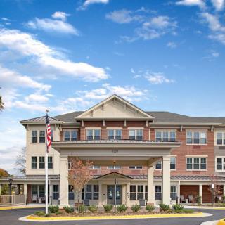 A three-story assisted living facility with a welcoming entrance and an American flag in front, set against a partly cloudy sky.