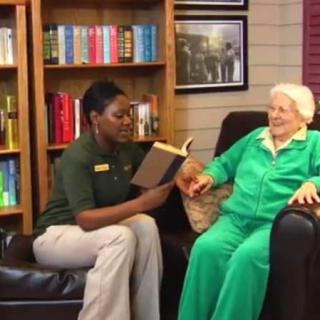 A caregiver reads a book to an elderly person in a cozy setting.
