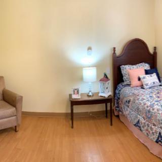 A cozy bedroom featuring a bed with decorative pillows, a nightstand with a lamp, and a comfortable chair on a wooden floor.