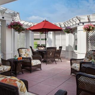 A cozy outdoor patio with wicker seating, a red umbrella, and hanging plants under a pergola on a sunny day.