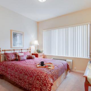 A cozy bedroom with a large bed covered in red bedding, illuminated by a table lamp next to a window with blinds.