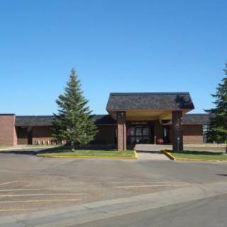 A school building with an entrance, surrounded by trees and a parking lot under a clear blue sky.