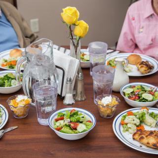 A neatly set dining table with plates of salad, main courses, water glasses, and yellow roses as a centerpiece.