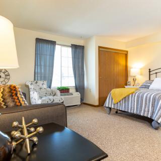 A cozy bedroom with stylish decor, featuring a bed, chair, and a large window letting in natural light.