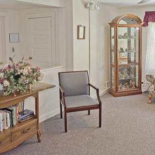 A cozy living room featuring a chair, wooden cabinet with glass doors, bookshelf, and floral decorations.