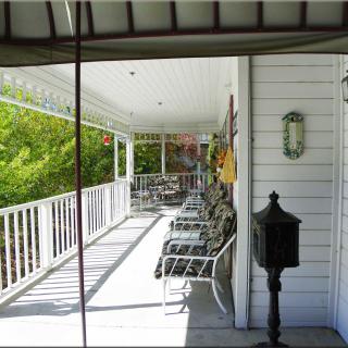 A sunny porch with several chairs lined up against a white wall, overlooking green foliage.