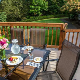 A patio deck setup with a dining table and chairs, featuring a meal and flowers overlooking a garden view.