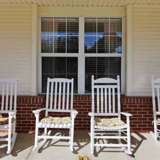 Four white rocking chairs sit on a porch in front of a window with brick siding.