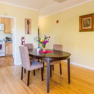 A bright dining room featuring a round table with chairs, flowers, and artwork on the wall, adjacent to a kitchen.