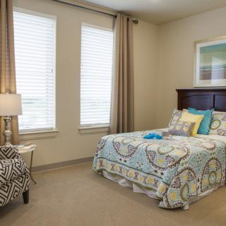 A cozy bedroom with a neatly made bed, decorated with patterned bedding, beside two windows with curtains and a lamp on the side table.