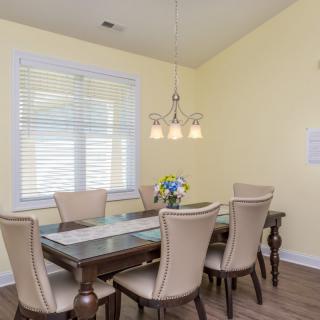 A bright dining room with a glass-top table and six upholstered chairs, under a decorative light fixture.