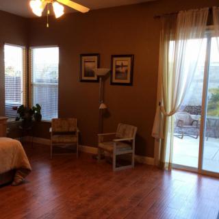 A cozy bedroom with wooden flooring, featuring two chairs, a bed, and large windows with curtains allowing natural light to fill the room.