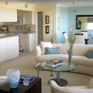 A modern living room with a beige sofa set, glass coffee table, and an open kitchen in the background.