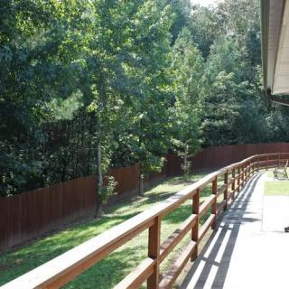 A sunny backyard with a wooden deck and railing overlooking trees and a fence.