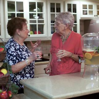 Two elderly women having a conversation in the kitchen while holding drinks.