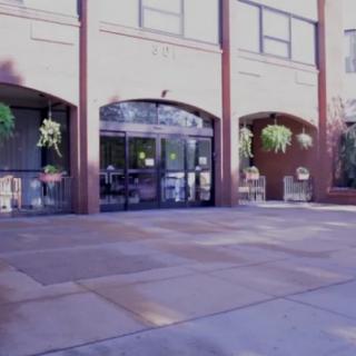 The entrance to a building with large windows and hanging plants, framed by brick arches.