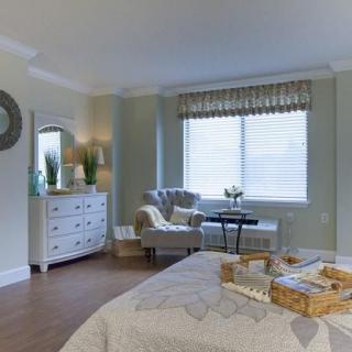 A cozy bedroom with a serene color palette and elegant furnishings, including an armchair, dresser, and window with soft natural lighting.