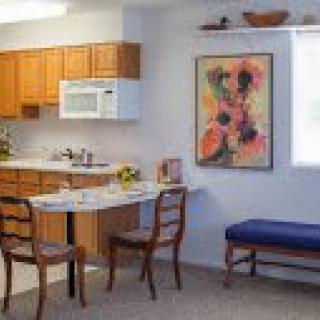 A cozy kitchen and dining area with wooden cabinets, a small table and chairs, and colorful artwork on the wall.