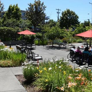 A scenic garden area with outdoor seating and umbrellas, surrounded by lush greenery and colorful flowers.