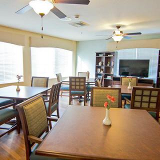 A well-lit dining room with multiple tables and chairs, featuring a television and bookshelf in the background.