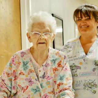 An elderly woman is assisted by a nurse in a healthcare setting.