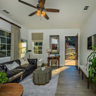 A cozy living room featuring a leather sofa, coffee table, ceiling fan, and large windows letting in natural light.