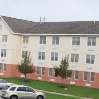 A residential apartment building with parked cars in the foreground and small trees on a grassy lawn.