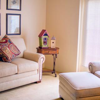 A cozy living room with a sofa, patterned cushions, and an ottoman beside a window. A small table holds colorful birdhouses as decor.