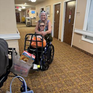 A caregiver assists an elderly individual in a wheelchair down a hallway.