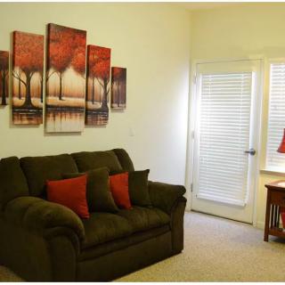 A cozy living room featuring a dark green sofa with red pillows, adorned with multi-panel wall art depicting trees. Natural light pours in through the window beside a small wooden table with a red lamp.