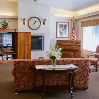 A cozy living room with a piano and floral decorations, featuring a large clock and comfortable seating.
