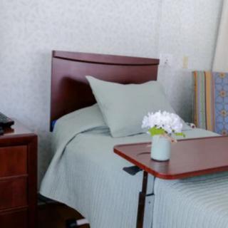 A hospital room with a neatly made bed, a nightstand, and a tray table with flowers.