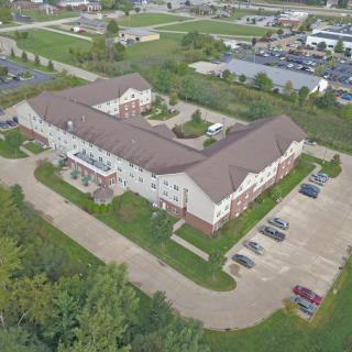 Aerial view of a large building with a parking lot surrounded by greenery and roads.
